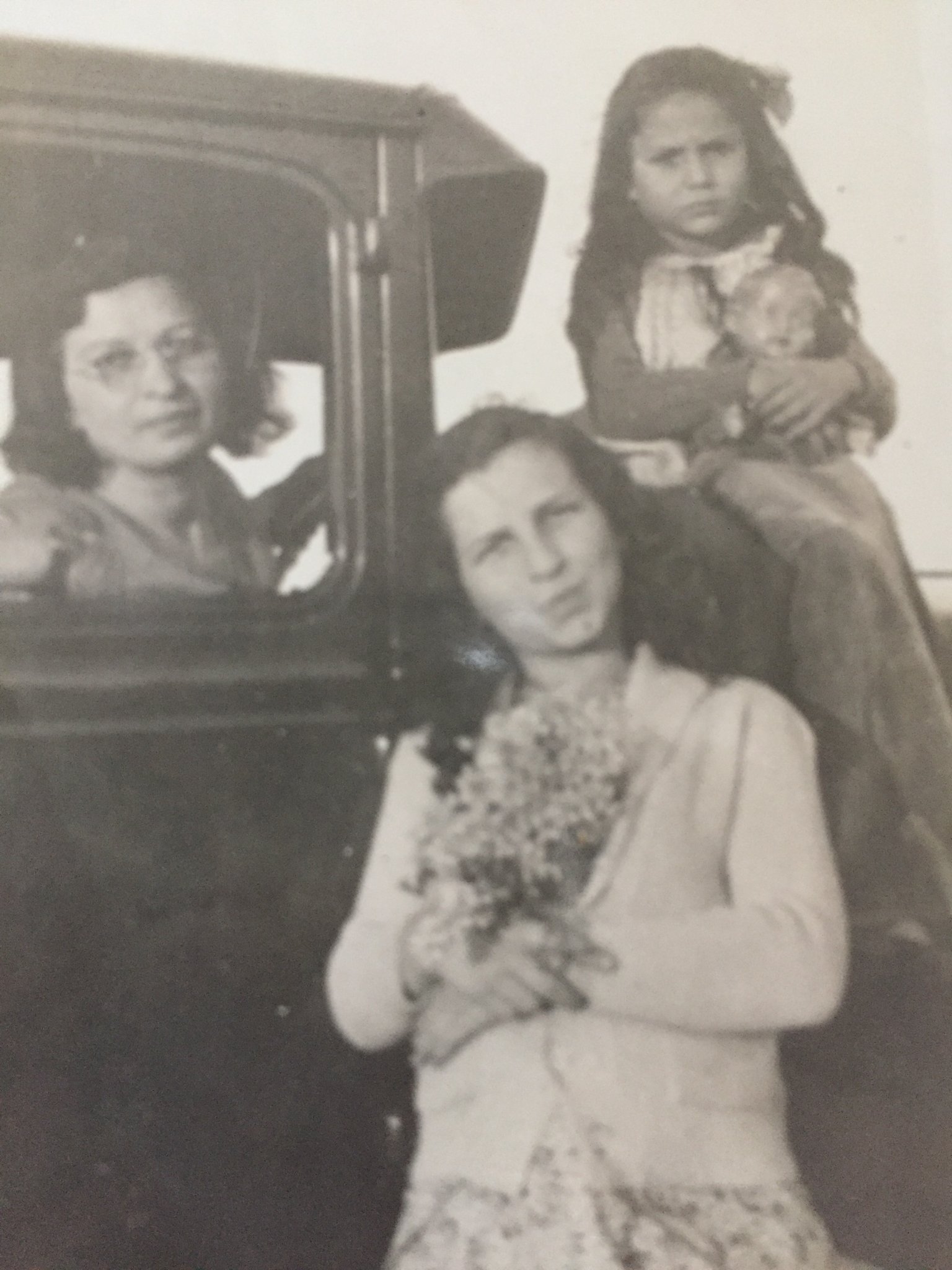 Kipp Dawson's mother sits on the hood of a 1930s car holding a doll. Her mother drives the car and her sister stands in front, holding a bunch of daisies.