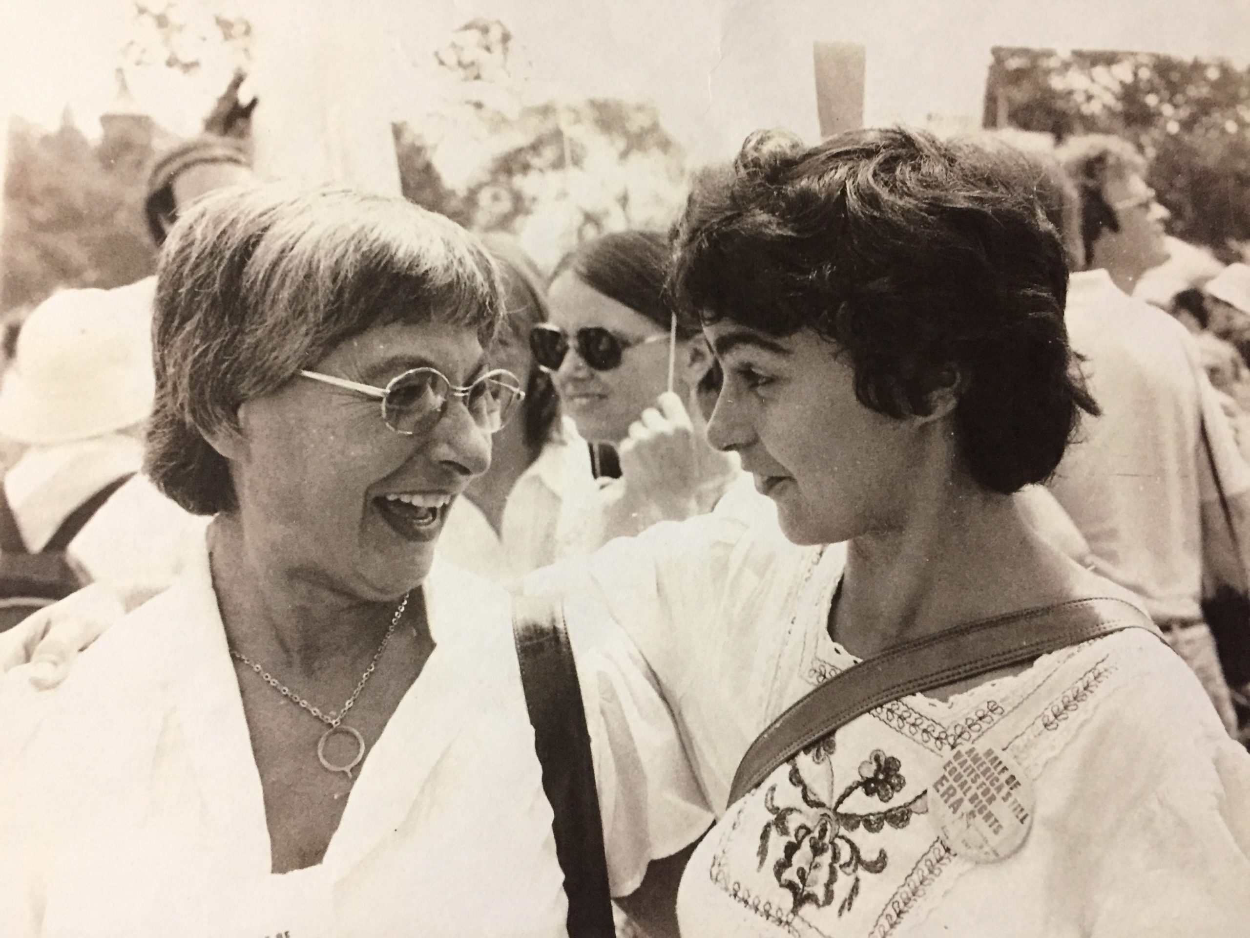 A young Kipp Dawson smiles and looks at her mother at a rally for women's rights. Dawson wears a button that says "half of America still waits for Equal Rights, ERA" and Kipp's mother wears a female symbol around her neck.