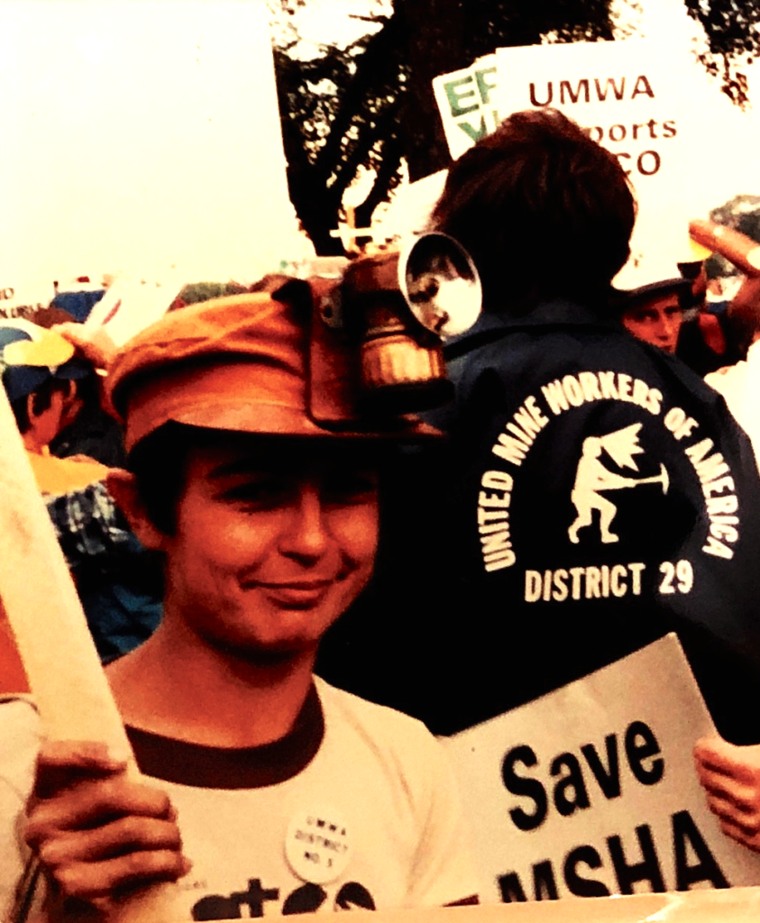 A colorized photo of Kipp Dawson holding a picket sign and wearing a miner's hat in front of a person's back that says "United Mine Workers of America, District 29"