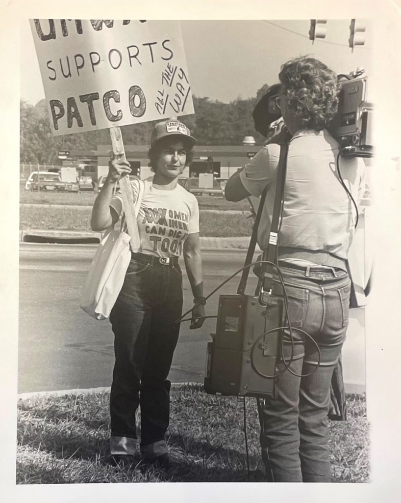 Kipp Dawson holds a picket sign that says "AMWA Supports PATCO all the way" and wears a shirt that says "Women Miners can dig it too!" A reporter with a video camera stands in front of her, shooting footage of Dawson.