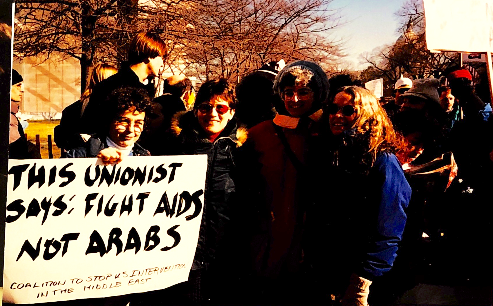 Kipp Dawson stands with a group of women at a gathering, holding a sign that says "This Unionist says: Fight AIDS, Not Arabs. Coalition to Stop US intervention in the Middle East"
