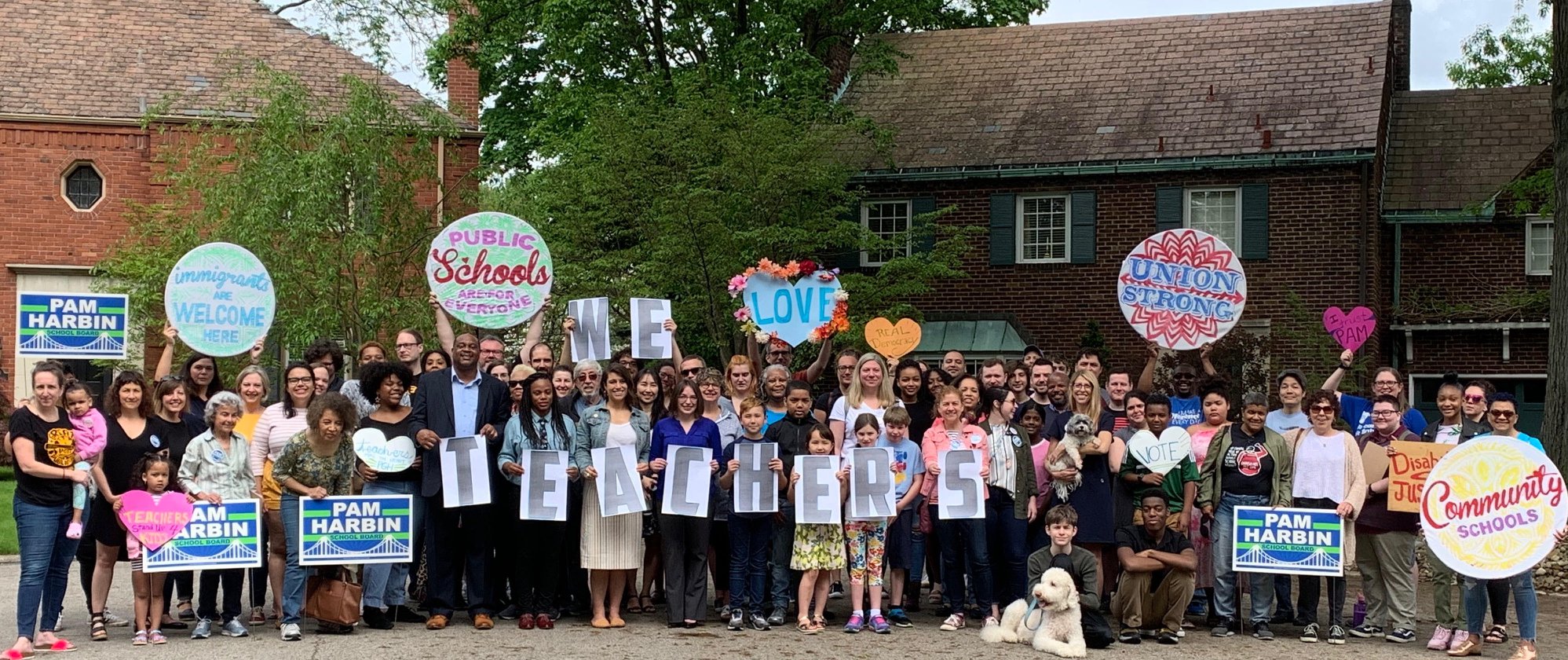 A diverse group of people gather with signs in support of public schools, candidate Pam Harbin, and a large sign held by many saying "WE LOVE TEACHERS"