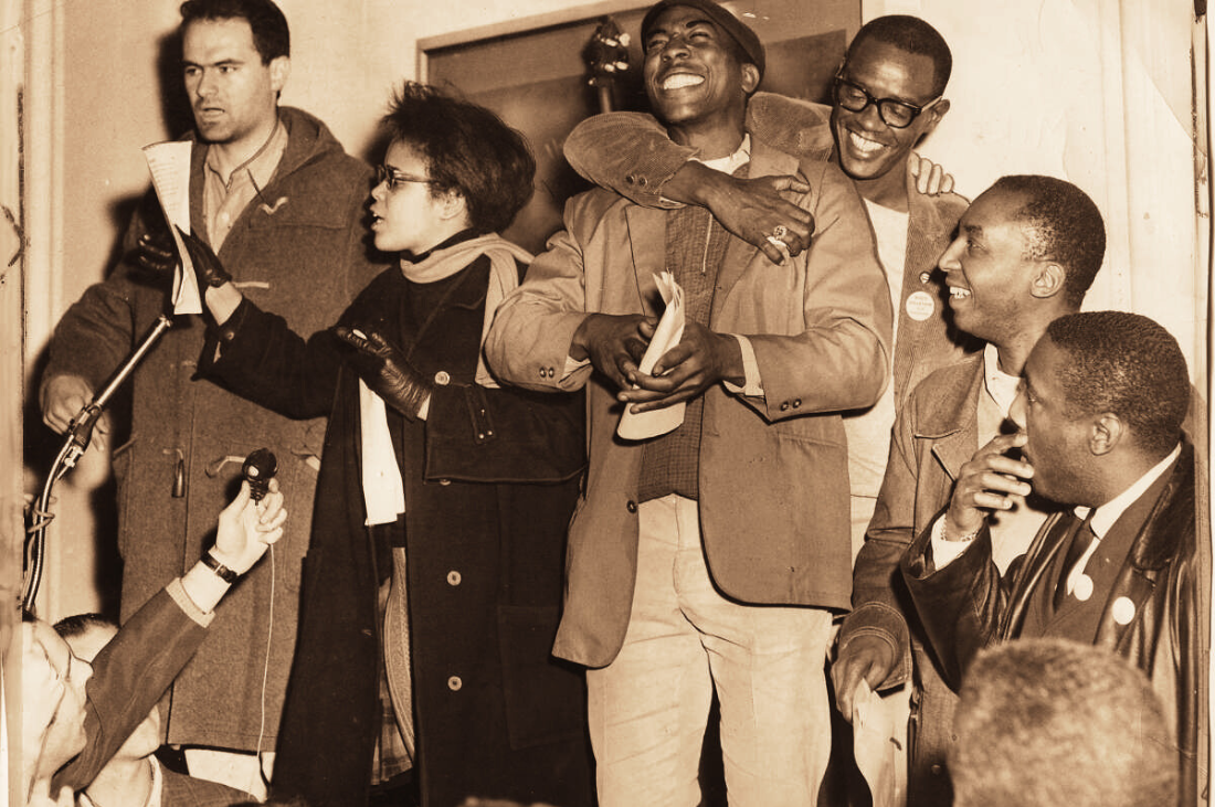 A sepia tone photograph of a group of men and women at a sit-in protest. One white man and a Black woman, Tracy Simms, are speaking into a microphone. Four Black men to the right of Tracy are laughing and embracing.